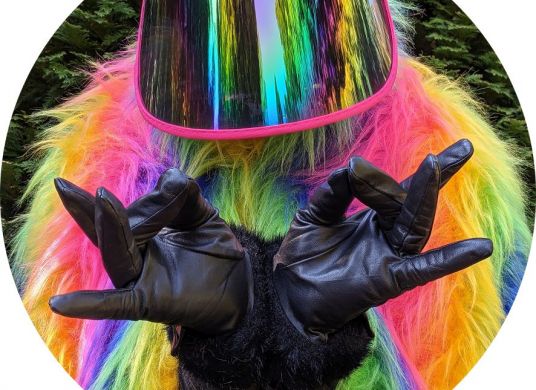 Hände in schwarzen Lederhandschuhen machen eine Geste vor einer verspiegelten Maske mit Fellkragen in Regenbogenfarben