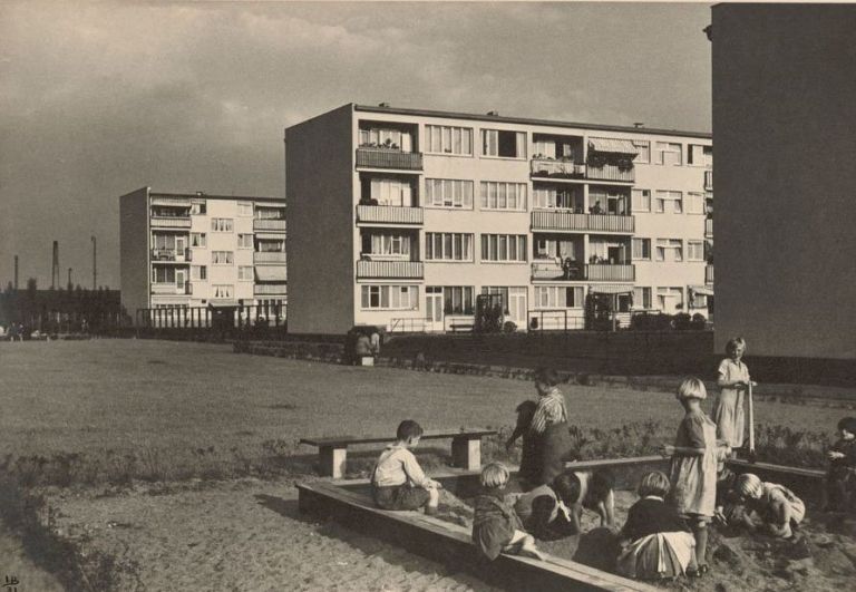 Schwarz-Weiß-Fotografie mit spielenden Kindern in einem Sandkasten im Vordergrund und Mehrfamilien-Siedlungshäusern im Hintergrund
